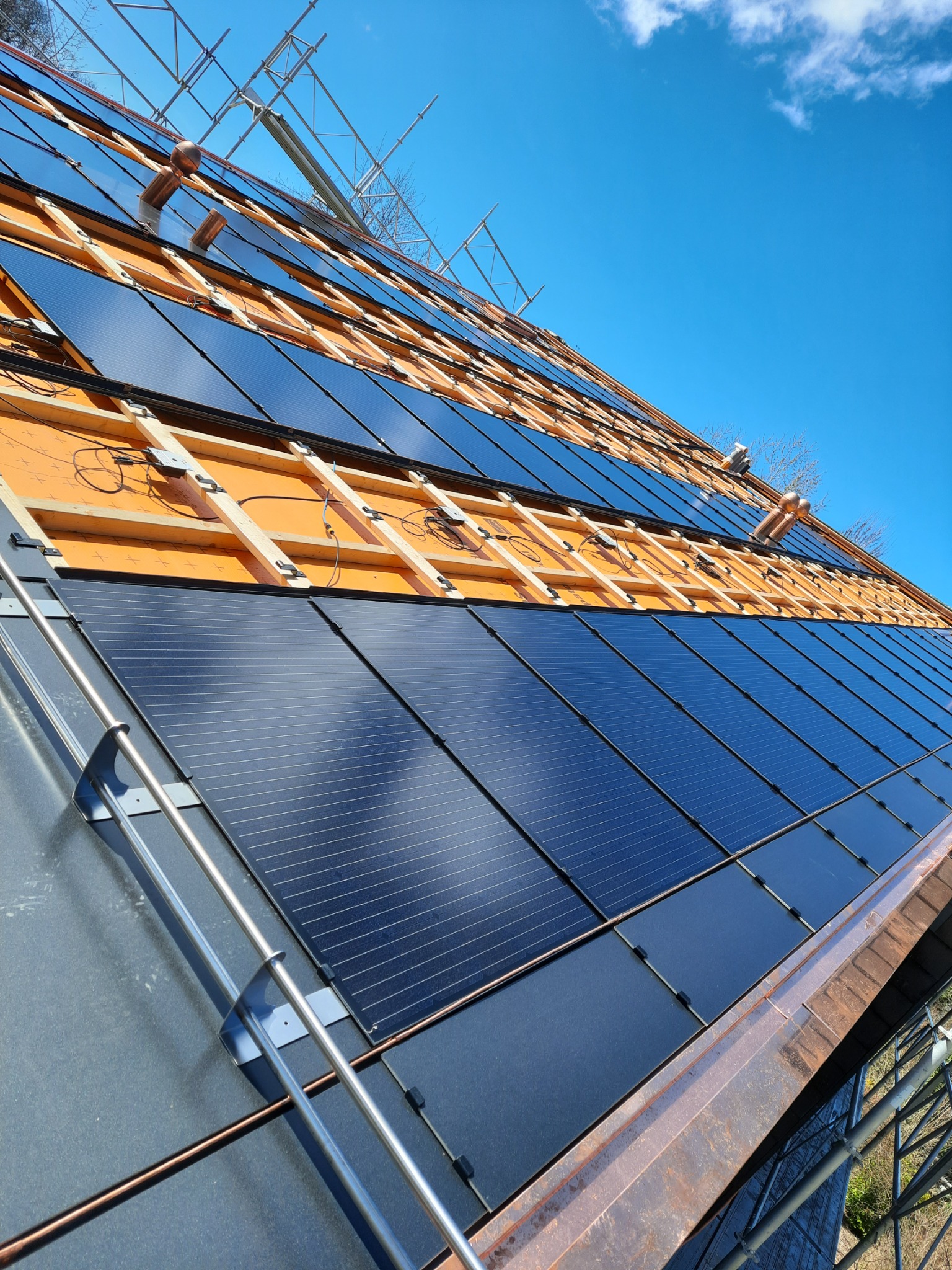 Montaż paneli fotowoltaicznych na dachu z widoczną konstrukcją drewnianą i metalowymi elementami montażowymi w słoneczny dzień.