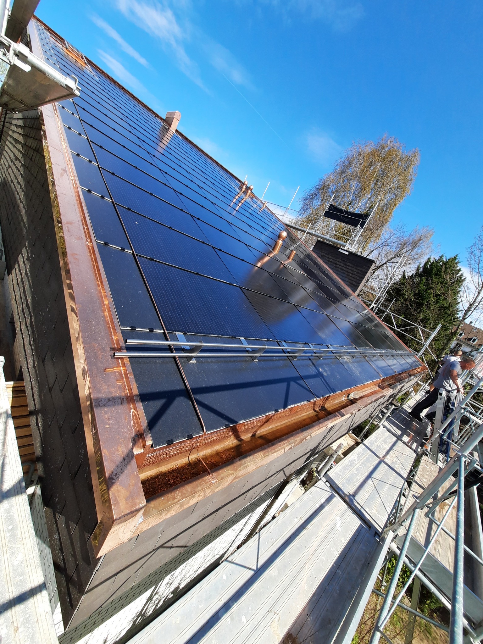 Montaż paneli słonecznych na dachu z miedzianymi rynnami, widok z rusztowania, pracownik w szarej koszulce na drabinie, błękitne niebo w tle.