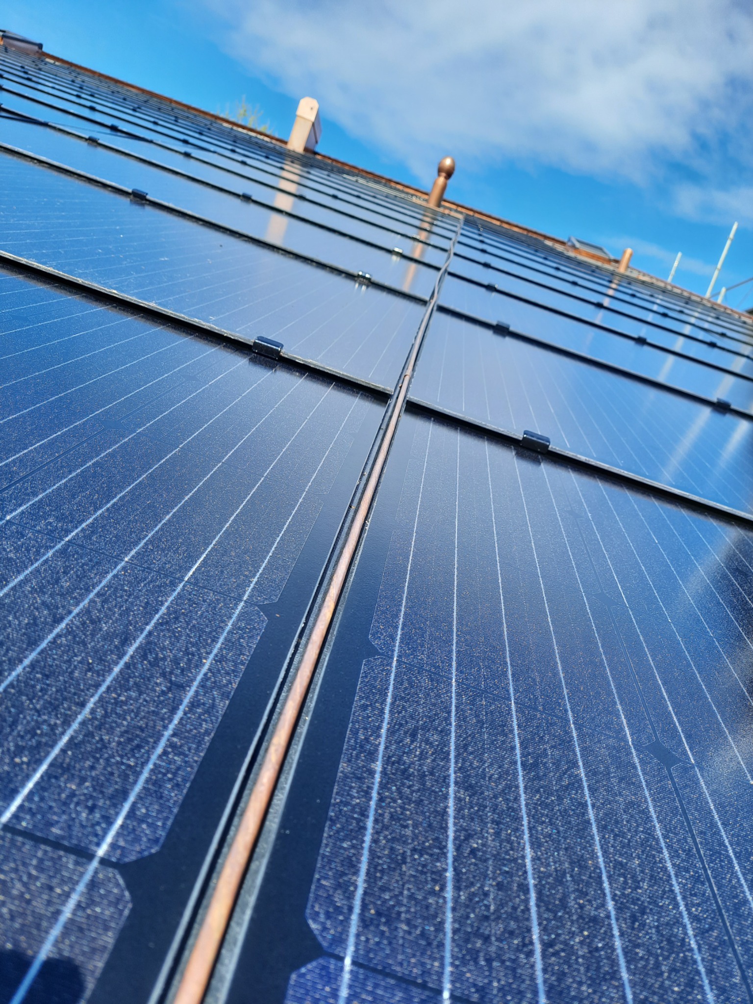 Instalacja paneli fotowoltaicznych na dachu z widocznymi elementami montażowymi i fragmentem komina na tle błękitnego nieba.