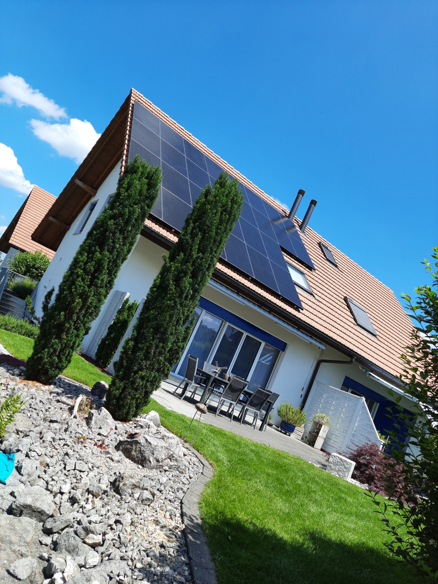 Nowoczesny dom jednorodzinny z panelami słonecznymi na dachu, widok z niskiej perspektywy, podkreślający instalację fotowoltaiczną i zadbany ogród z kamiennym skalniakiem na pierwszym planie.