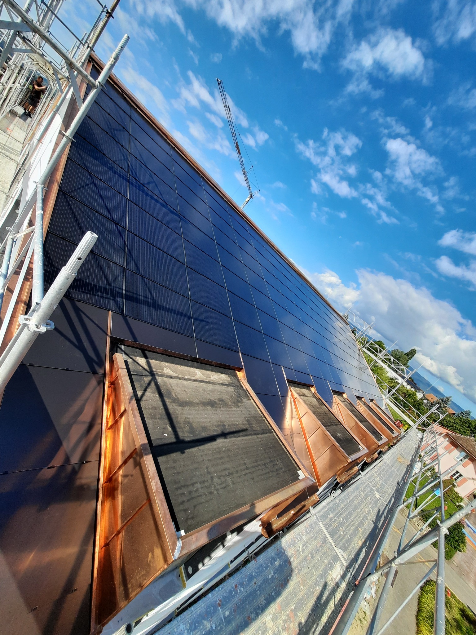 Instalacja paneli fotowoltaicznych na dachu z miedzianymi elementami wykończeniowymi, widoczne rusztowania i żuraw budowlany w tle, błękitne niebo z chmurami.