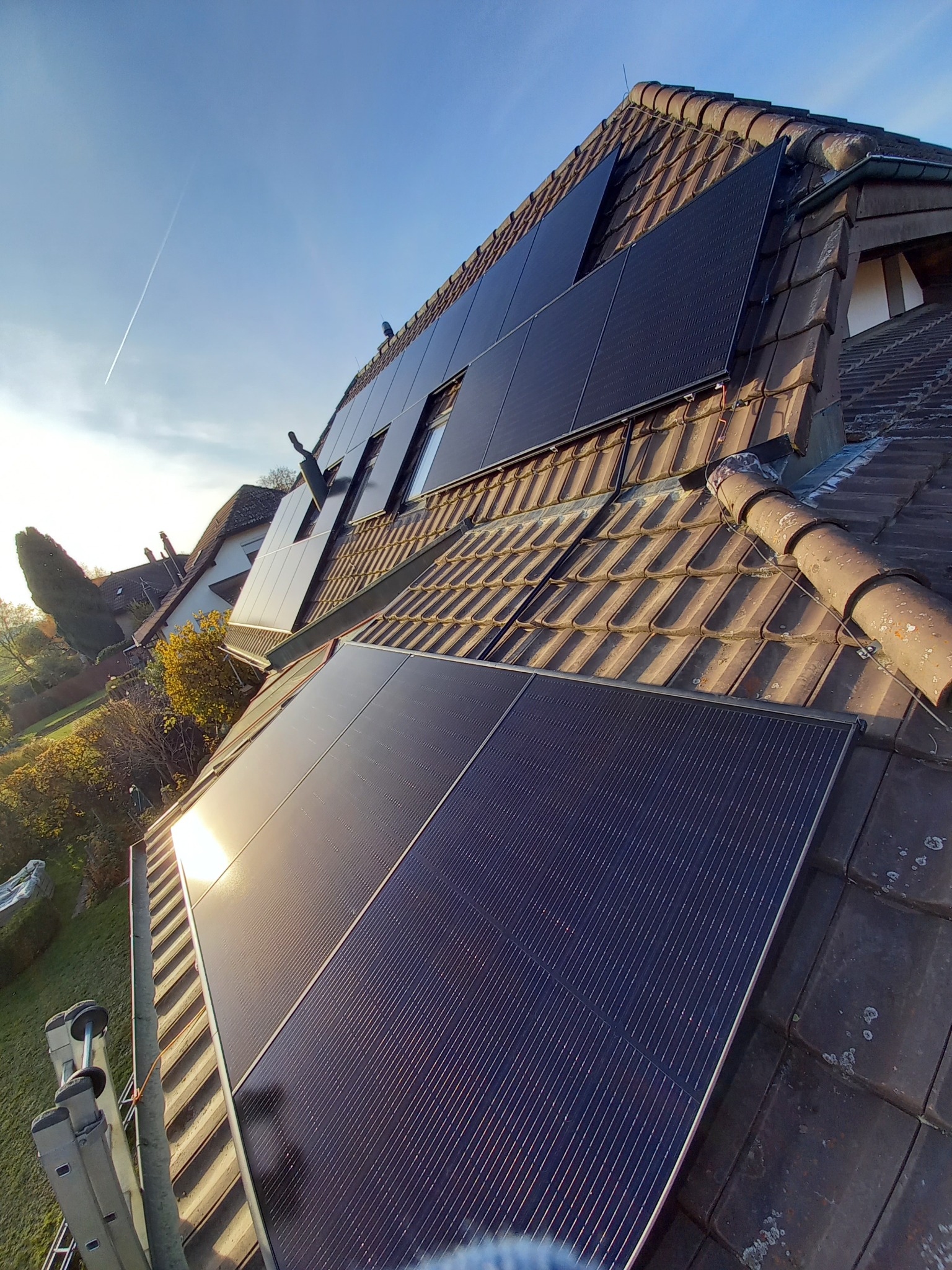 Montaż paneli fotowoltaicznych na brązowym, ceramicznym dachu domu jednorodzinnego, widok z drabiny, słoneczny dzień.