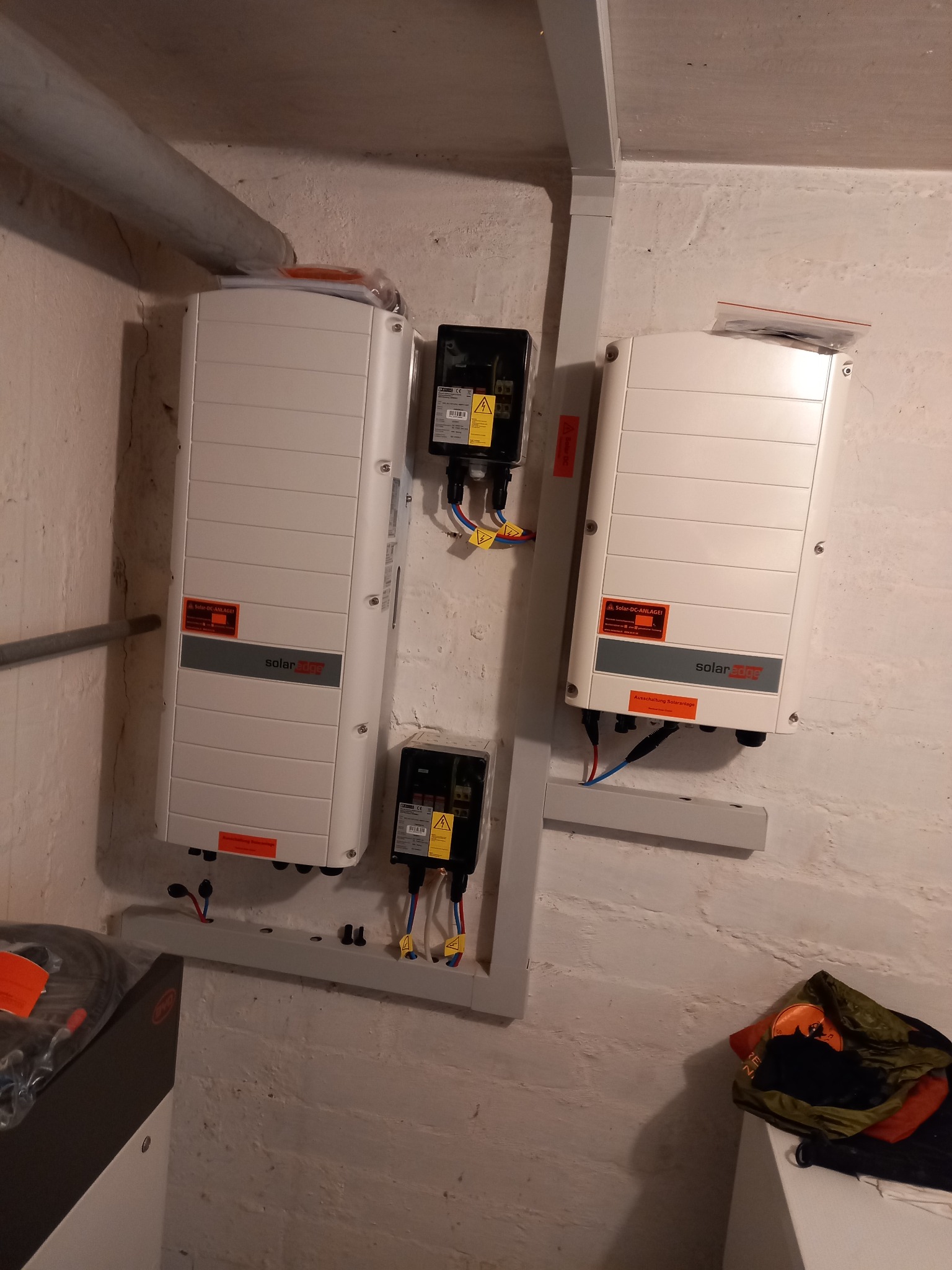 Dwa białe falowniki SolarEdge zamontowane na ścianie z widocznymi przewodami, czarna skrzynka elektryczna, rura instalacyjna i akumulator w tle.