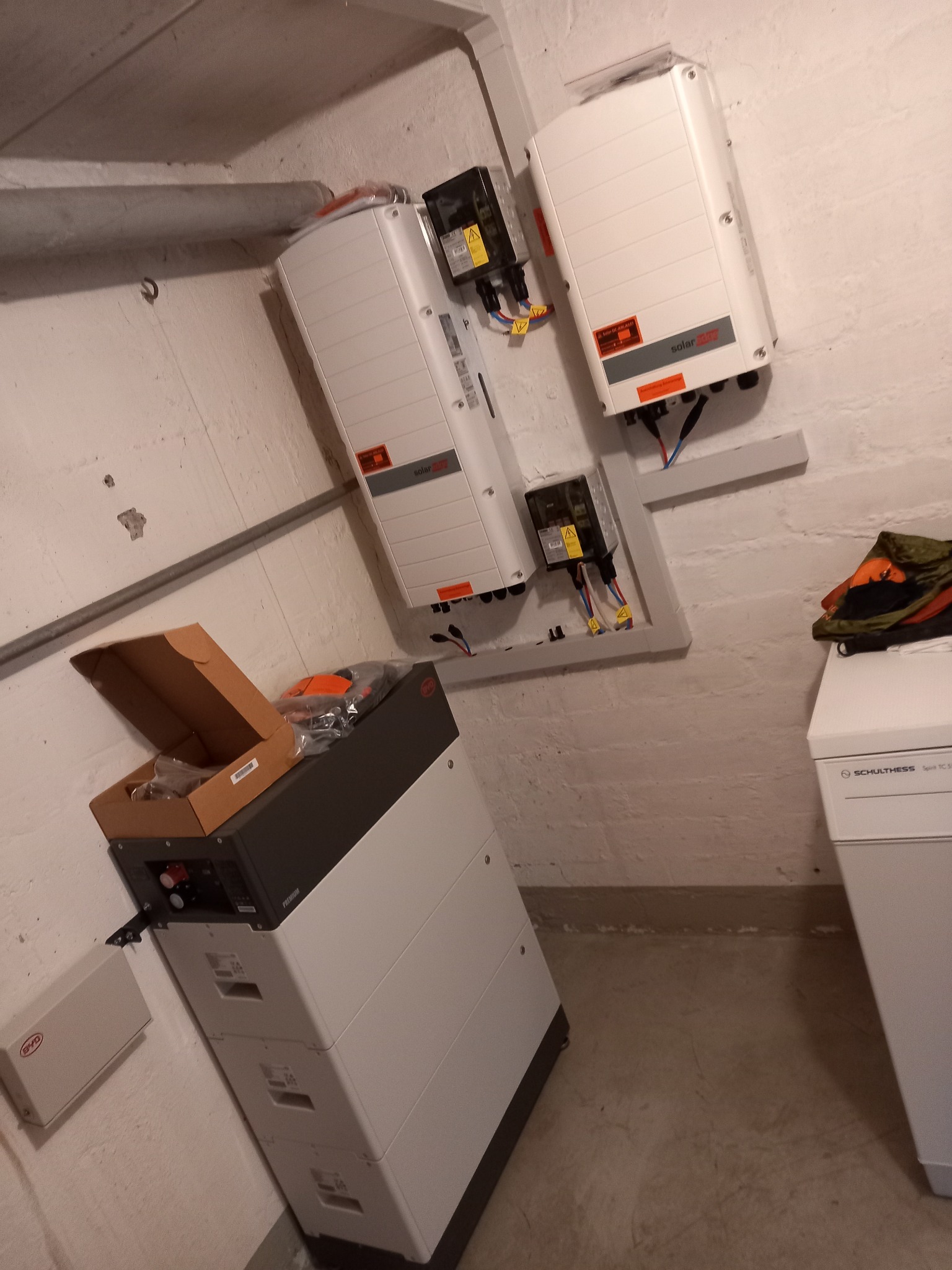 Elementy instalacji fotowoltaicznej w piwnicy: dwa inwertery SolarEdge zamontowane na ścianie, obok akumulator BYD z otwartym kartonem, widoczne przewody i rury.