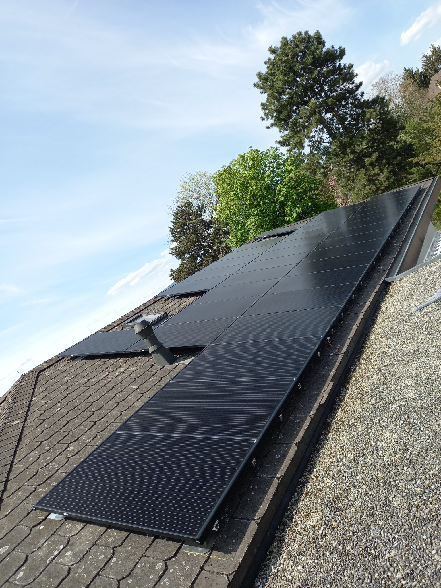 Czarne panele słoneczne zamontowane na spadzistym dachu pokrytym szarymi dachówkami, widoczne drzewa i błękitne niebo w tle.