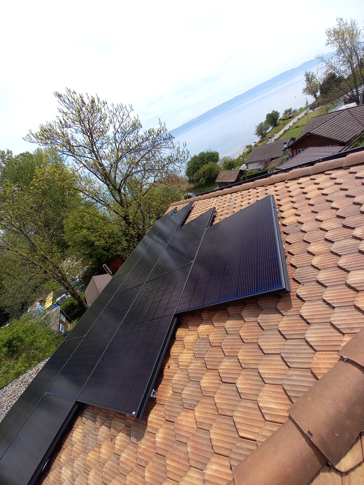Częściowo zamontowane czarne panele słoneczne na dachu z dachówki ceramicznej, widok na jezioro i zabudowania w tle.
