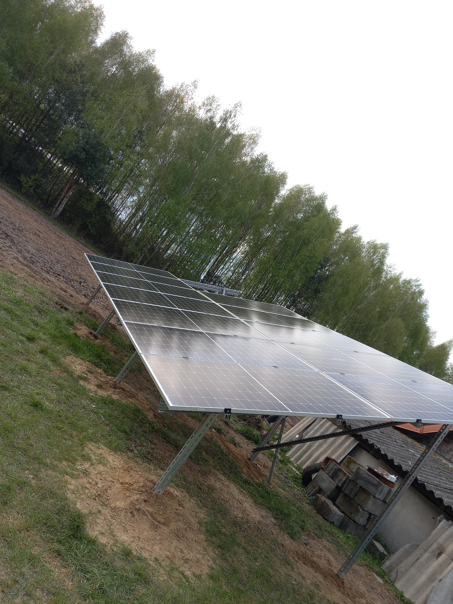 Panel słoneczny zamontowany na metalowej konstrukcji na skraju pola z widokiem na las, obok budynku gospodarczego z szarym dachem. Widoczne odbicie nieba w panelach.