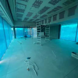 Wnętrze w trakcie remontu z rozstawionymi rusztowaniami, widoczne przygotowane kable elektryczne i częściowo wykończony sufit z widocznymi śladami po szpachlowaniu. Okna zabezpieczone folią. Całość...