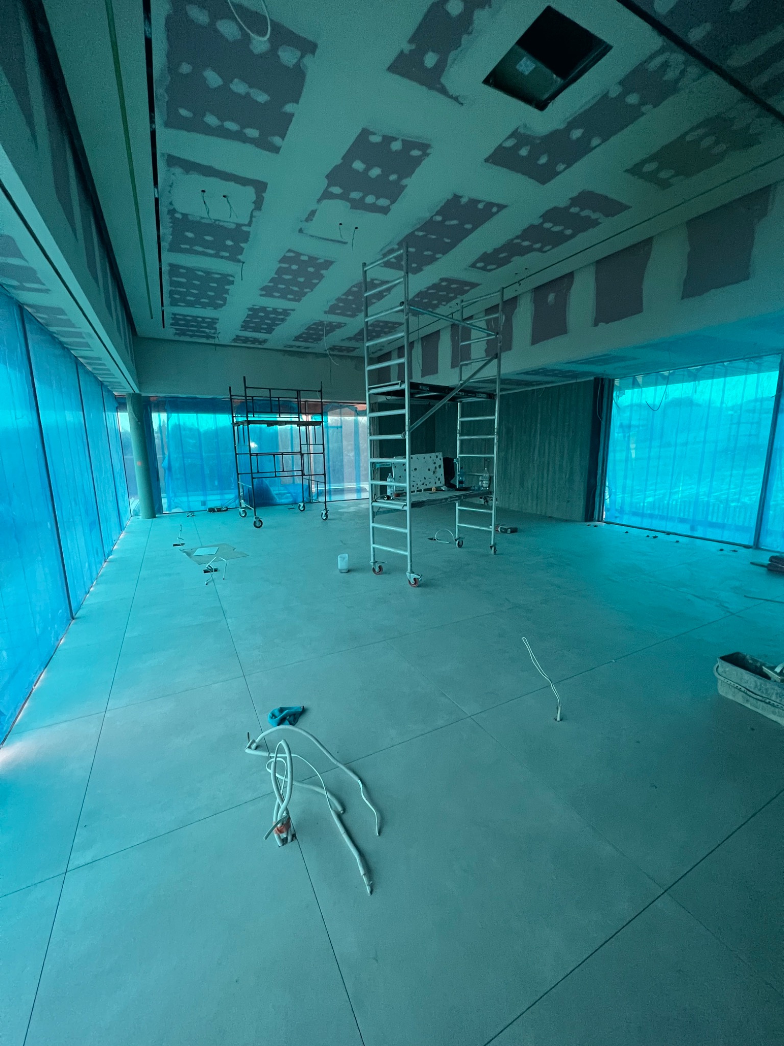 Wnętrze w trakcie remontu z rozstawionymi rusztowaniami, widoczne przygotowane kable elektryczne i częściowo wykończony sufit z widocznymi śladami po szpachlowaniu. Okna zabezpieczone folią. Całość...