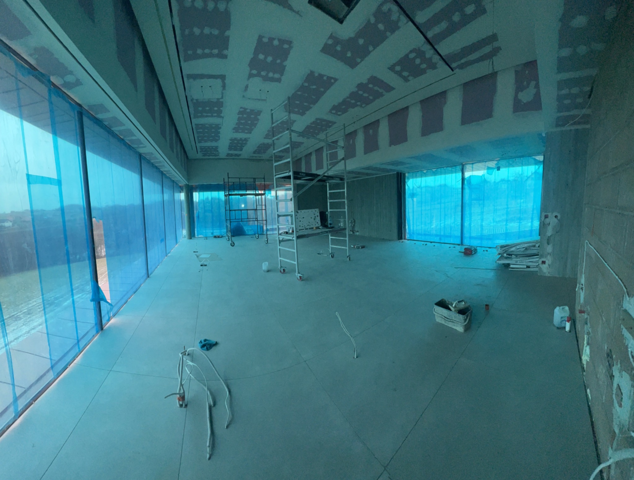 Wnętrze w trakcie szpachlowania sufitu z widocznymi rusztowaniami, zabezpieczonymi oknami folią i elementami instalacji wystającymi z podłogi.