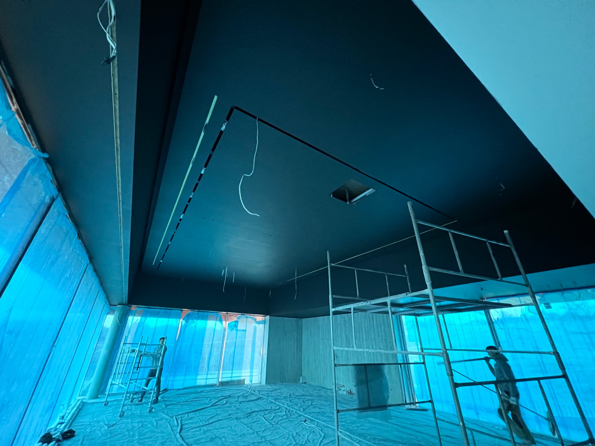 Wnętrze w trakcie prac wykończeniowych: czarny sufit z wystającymi przewodami, ściany przygotowane do malowania, podłoga zabezpieczona folią, widoczne rusztowania i sylwetki dwóch osób.