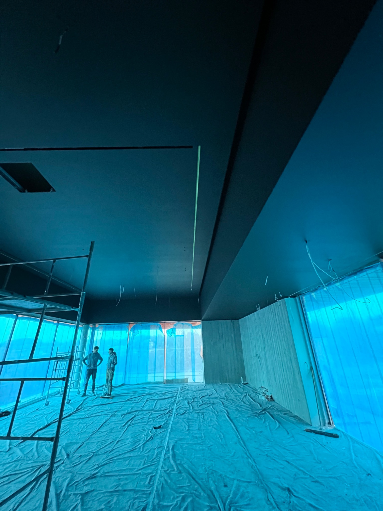 Wnętrze w trakcie prac wykończeniowych: pomalowany na czarno sufit, zabezpieczona folią podłoga, widoczne rusztowanie i dwie osoby w oddali.