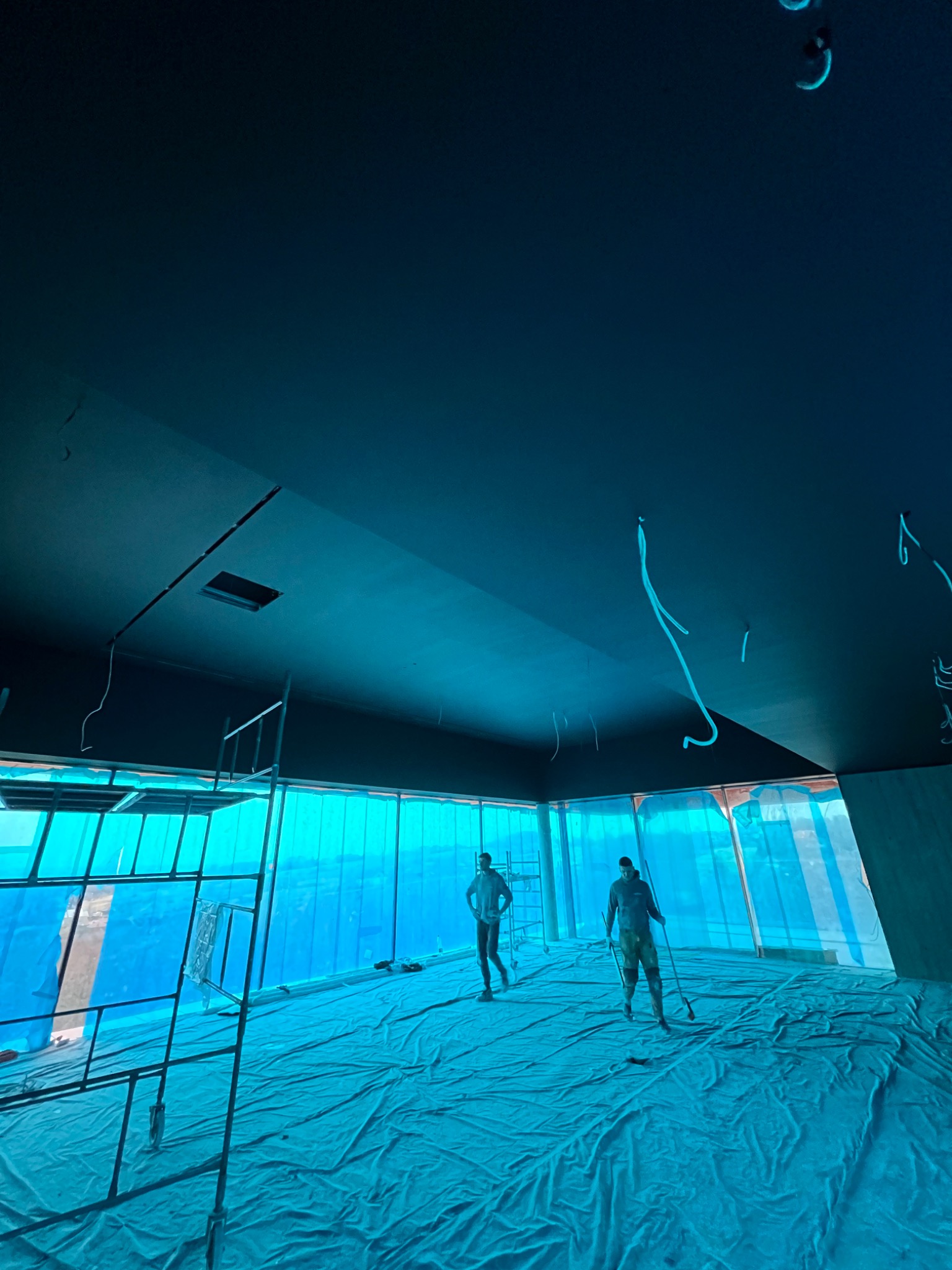 Wnętrze w trakcie prac wykończeniowych: dwóch pracowników na zabezpieczonej folią podłodze, jeden z narzędziem do rozprowadzania masy na długim kiju, sufit pokryty świeżą warstwą wykończeniową...