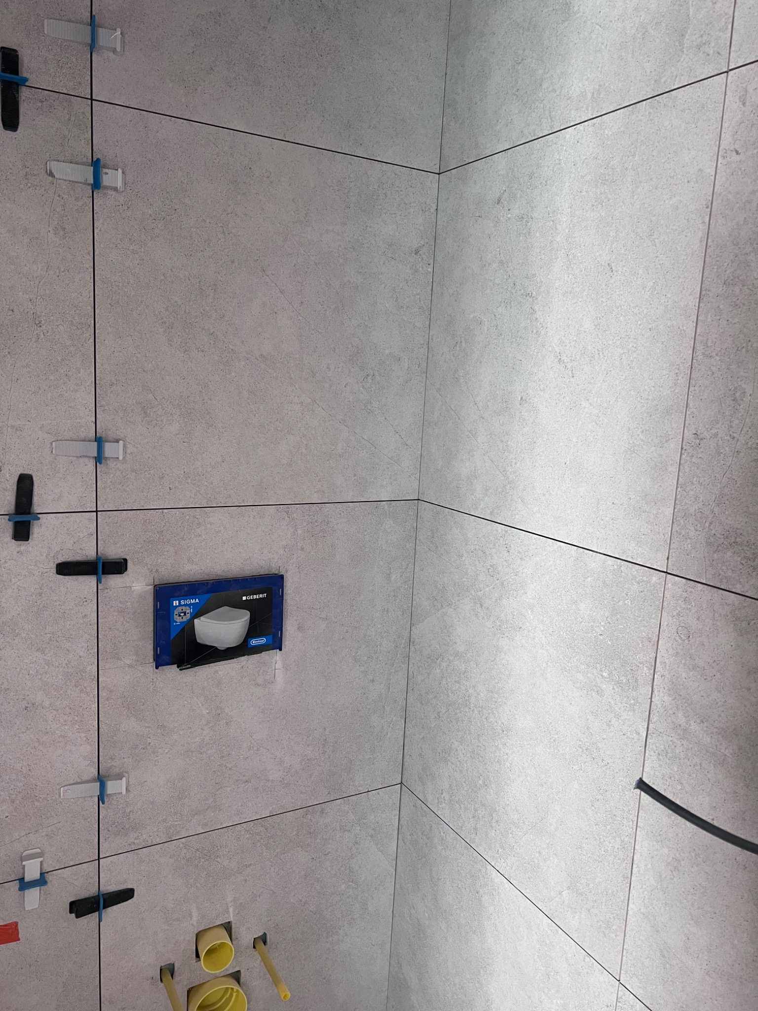 Świeżo ułożone, szare płytki ścienne z krzyżykami dystansowymi, widoczne żółte rury instalacji sanitarnej i szablon do montażu podtynkowego zestawu WC.