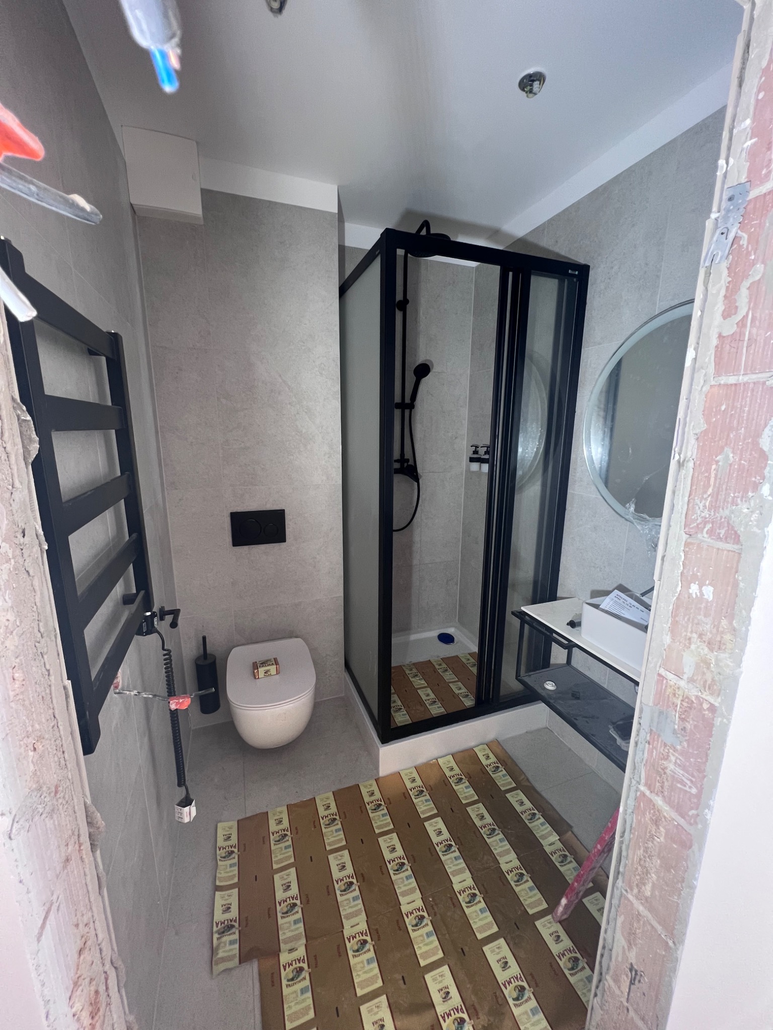 Łazienka w trakcie remontu: widoczne płytki podłogowe zabezpieczone kartonem, kabina prysznicowa z czarnymi ramami, grzejnik drabinkowy, toaleta i umywalka z lustrem. Fragment nieotynkowanej ściany...