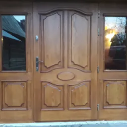 Drewniane drzwi zewnętrzne dwuskrzydłowe z bocznymi naświetlami, widoczne detale frezowania i naturalne usłojenie drewna, w szybach odbicie otoczenia.