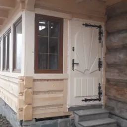 Wejście do drewnianego domu z oknem i drzwiami zdobionymi czarnymi, kutymi zawiasami, widoczne betonowe schody i kamienny fundament.