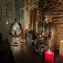 Ustawienie w gabinecie: statuetka Buddy trzymająca zapaloną świeczkę, ozdobny lampion, zapalone świece, karty tarota na drewnianym blacie, ściana z kamienia dekoracyjnego, certyfikaty na ścianie...