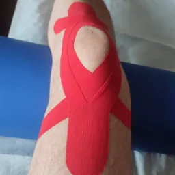 Noga z aplikacją czerwonej taśmy kinesiotaping na kolanie, ułożona na białym podkładzie i niebieskim wałku, widok z góry.