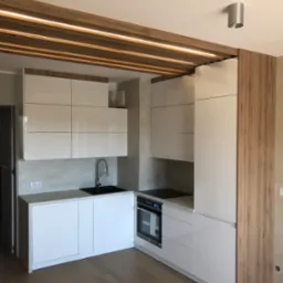 Nowoczesna kuchnia z białymi, lakierowanymi frontami szafek, czarnym zlewem i drewnianym akcentem na suficie z oświetleniem LED.