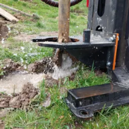 Wiercenie studni w Kraśniku: woda tryska spod wiertła, błoto i trawa wokół, fragment maszyny wiertniczej z pomarańczowym elementem mocującym.