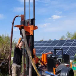 Pracownik w pomarańczowych rękawicach obsługuje wiertnicę studni głębinowej na tle paneli słonecznych i błękitnego nieba.