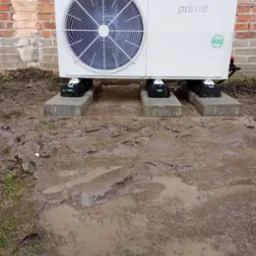 Biała pompa ciepła marki Prima, model z czynnikiem chłodniczym R32, umieszczona na betonowych bloczkach, na tle ceglanej ściany i błotnistego terenu.