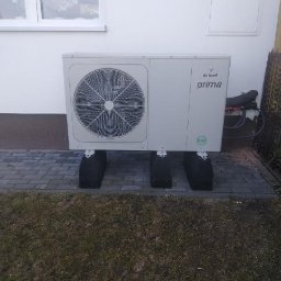 INSTALACJE CHROSTOWSKI Marcin Chrostowski - Biała pompa ciepła Brötje Prima, zamontowana na czarnych podstawach, na tle białej ściany budynku z oknem. Widoczny fragment trawnika i kostki brukowej.