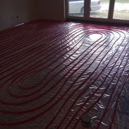 Rozłożona instalacja ogrzewania podłogowego z czerwonych rur na folii z siatką, widok z góry, w tle okno z widokiem na samochód.