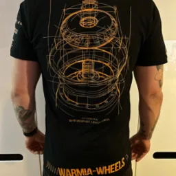 Czarna koszulka z pomarańczowym technicznym rysunkiem koła zamachowego i napisem 'WARMIA-WHEELS' na dole, widok z tyłu na mężczyznę z tatuażami na ramionach.