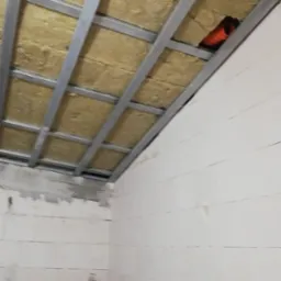 Poddasze w trakcie ocieplania, widoczna wełna mineralna między metalowymi profilami konstrukcyjnymi, z pomarańczową rurą wentylacyjną w izolacji, ściany z białych bloczków.