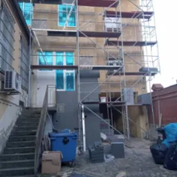 Budynek w rusztowaniach podczas prac ociepleniowych, okna zabezpieczone folią, widoczny fragment ocieplonej ściany styropianem, worki z klejem, oraz narzędzia budowlane na ziemi.