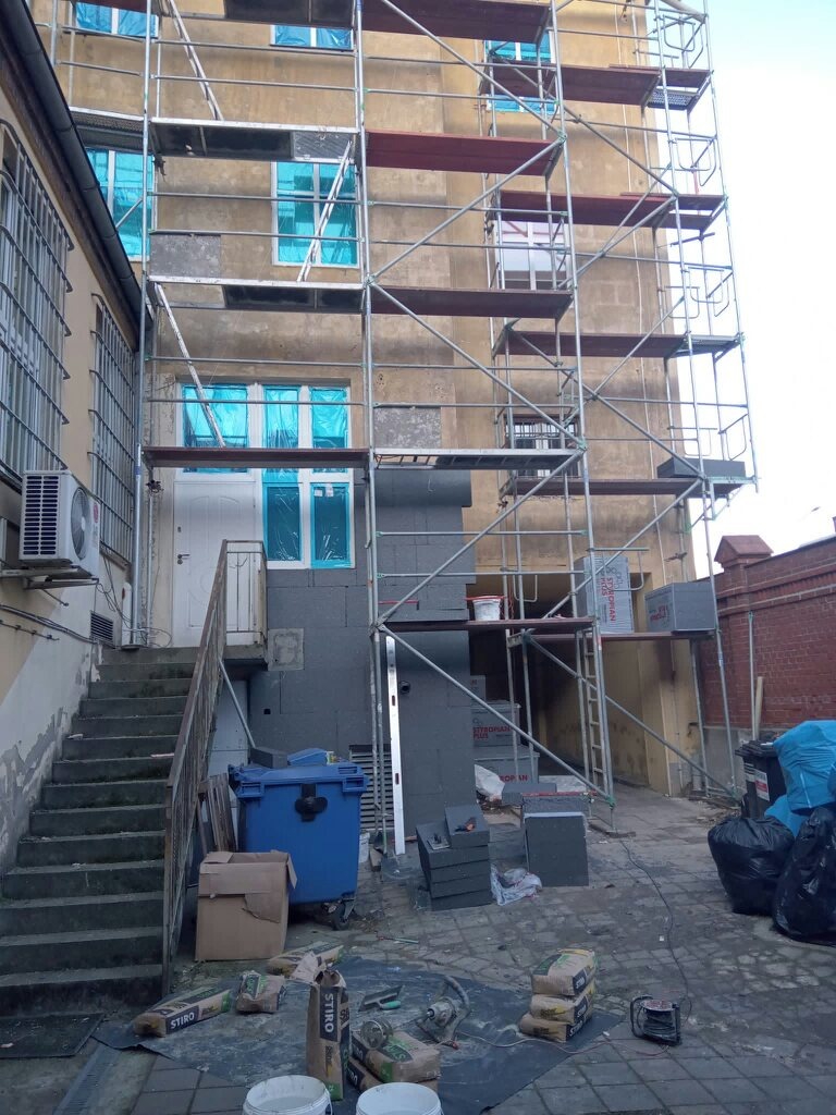 Budynek w rusztowaniach podczas prac ociepleniowych, okna zabezpieczone folią, widoczny fragment ocieplonej ściany styropianem, worki z klejem, oraz narzędzia budowlane na ziemi.