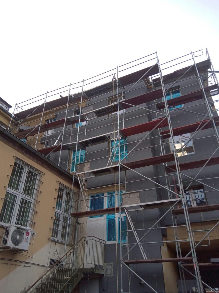 Budynek w trakcie ocieplania, widoczne rusztowania, zabezpieczone okna folią, szare płyty izolacyjne na elewacji, klimatyzator na ścianie sąsiedniego budynku.