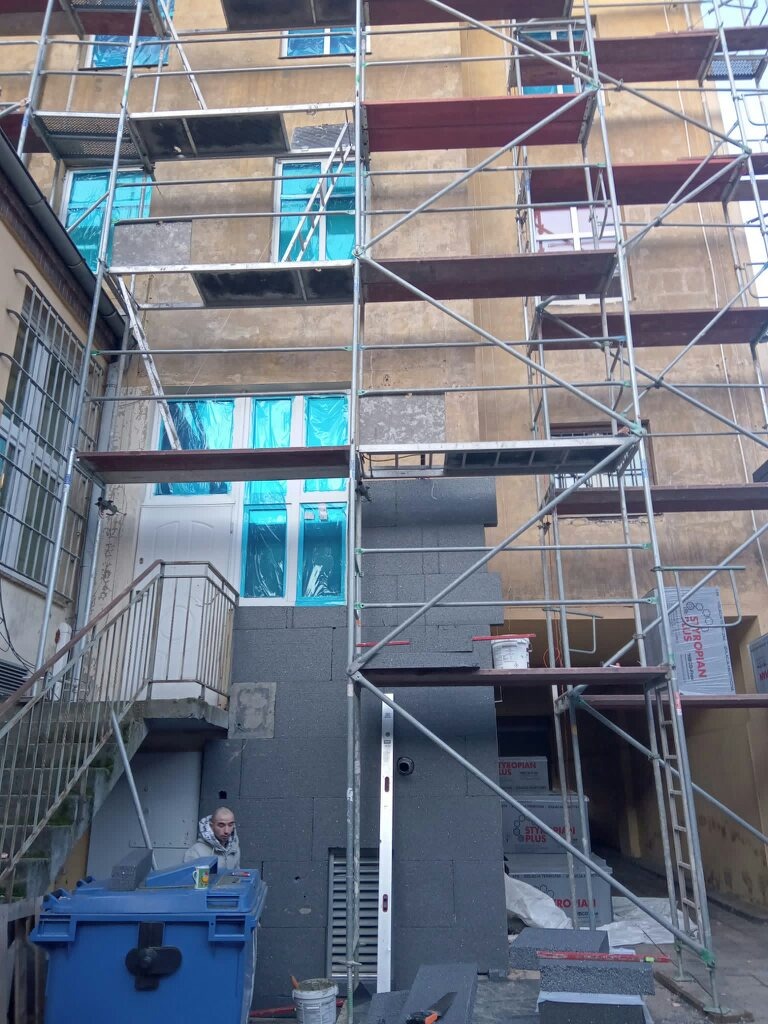 Budynek w trakcie prac: fragment ocieplonej ściany z szarym styropianem, rusztowanie, okna zabezpieczone folią, pracownik w dole kadru z kubkiem w ręku.