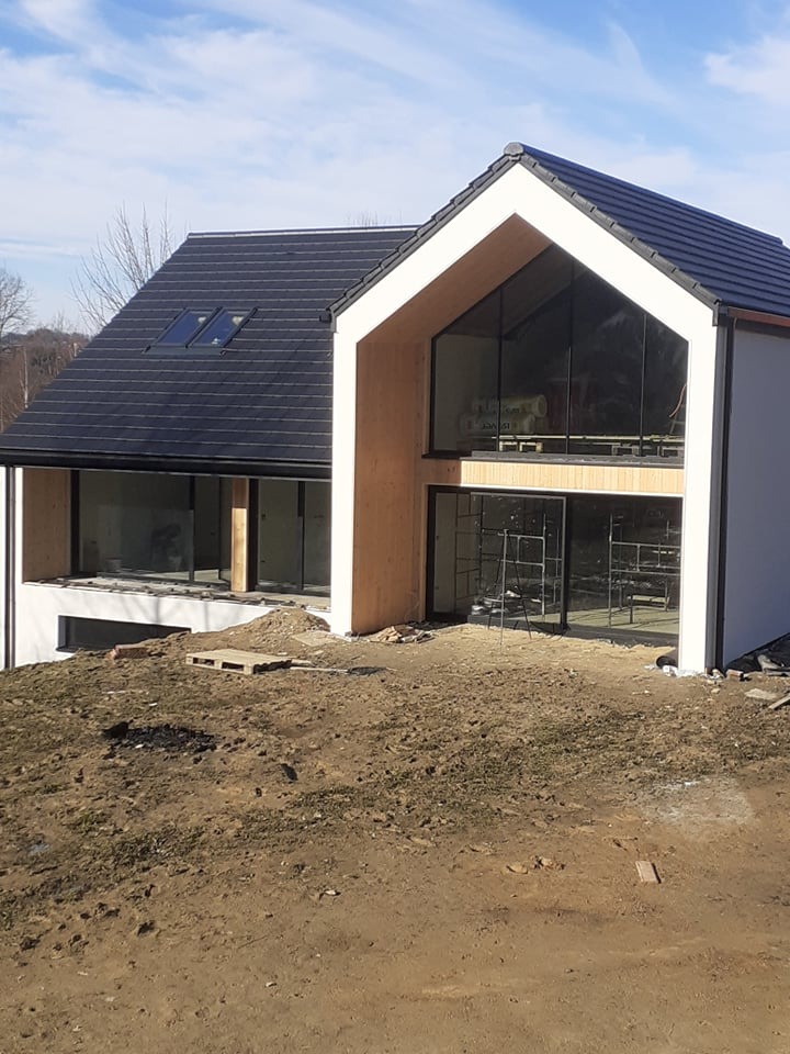 Nowoczesny dom jednorodzinny w trakcie budowy, z widocznymi dużymi oknami, czarną dachówką i surowym otoczeniem z ziemią i paletą.