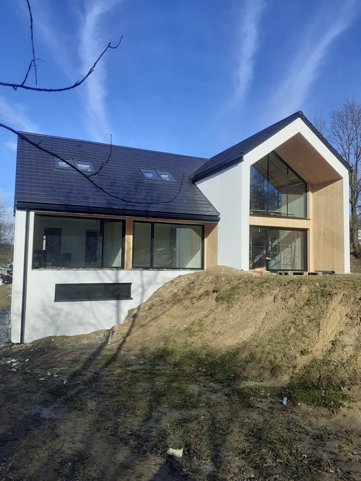 Nowoczesny dom jednorodzinny z białymi ścianami i drewnianą elewacją, czarny dach z oknami dachowymi, widoczny nasyp ziemi przed budynkiem, błękitne niebo.