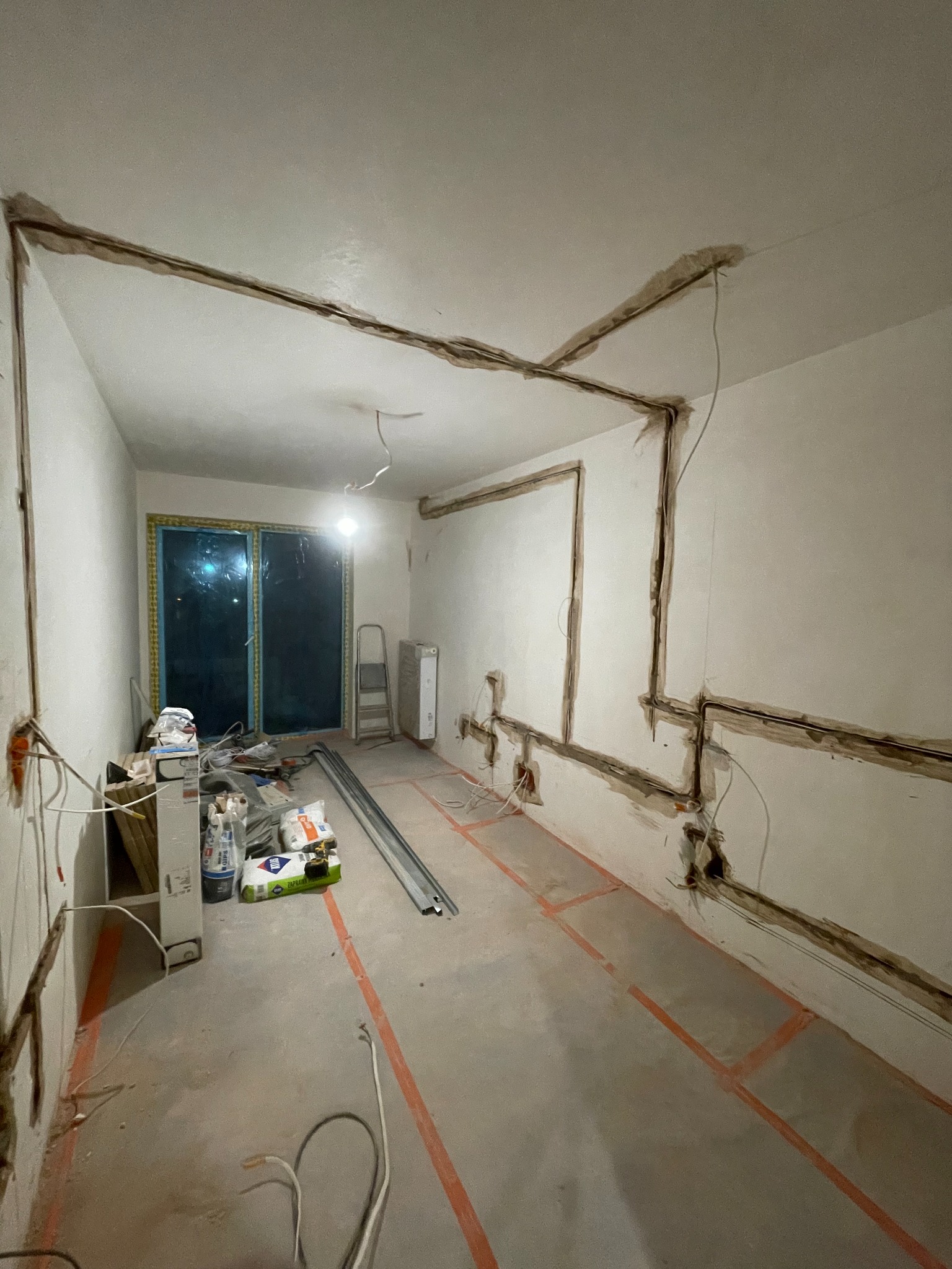 Wnętrze w trakcie prac instalacyjnych: wyfrezowane bruzdy pod przewody elektryczne na ścianach i suficie, widoczne puszki elektryczne, okno zabezpieczone folią, materiały budowlane na podłodze.