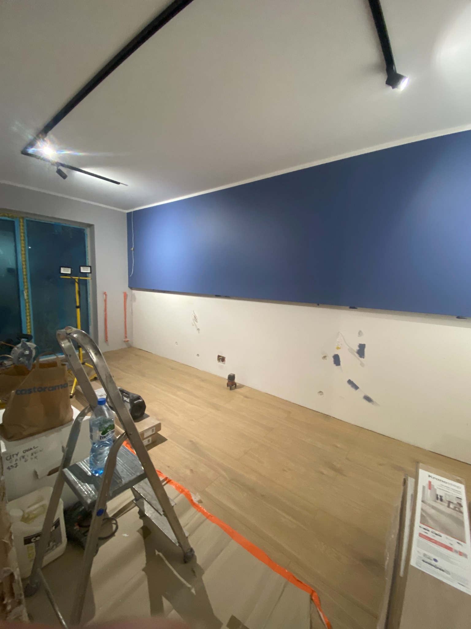 Pokój w trakcie prac wykończeniowych: pomalowana na granatowo górna część ściany kontrastuje z białą dolną, widoczne ślady po szpachlowaniu, ułożona drewniana podłoga, drabina, worki i narzędzia...