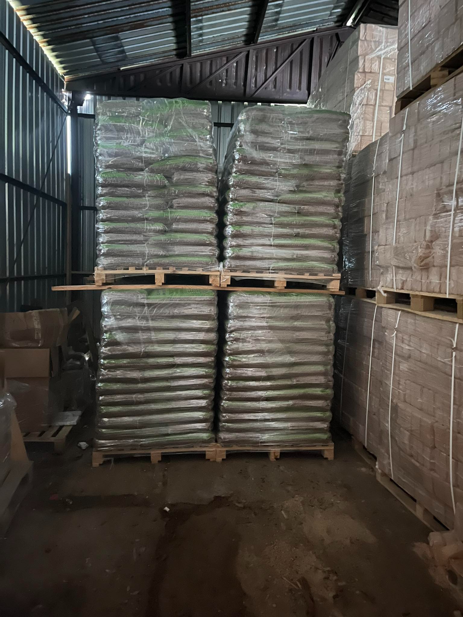 Magazyn pelletu: Cztery palety pelletu w przezroczystej folii, ułożone jedna na drugiej, w ciemnym magazynie z blachy falistej.