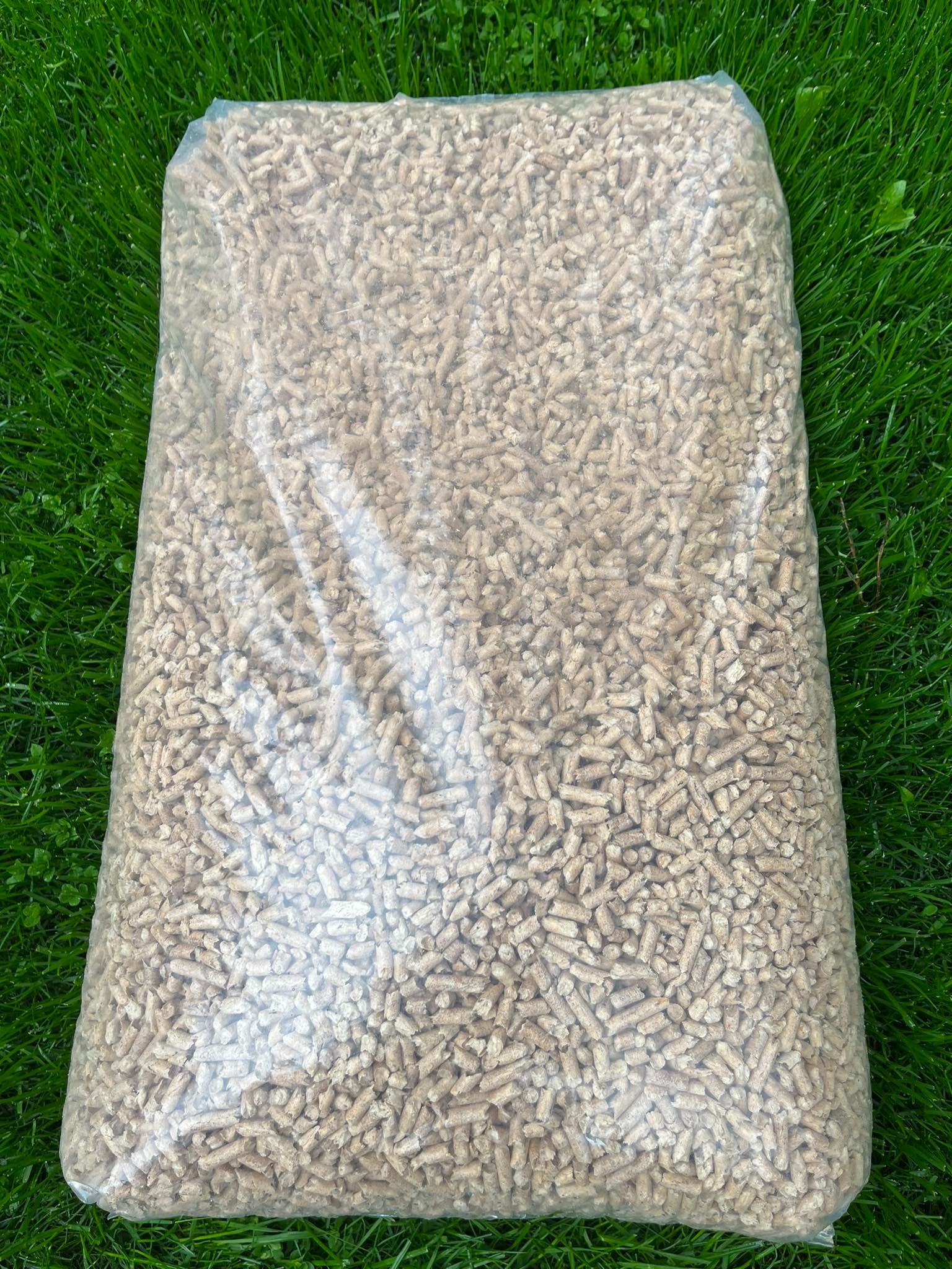 Worek przezroczysty wypełniony jasnymi, drewnianymi pelletami, leżący na soczystej, zielonej trawie.