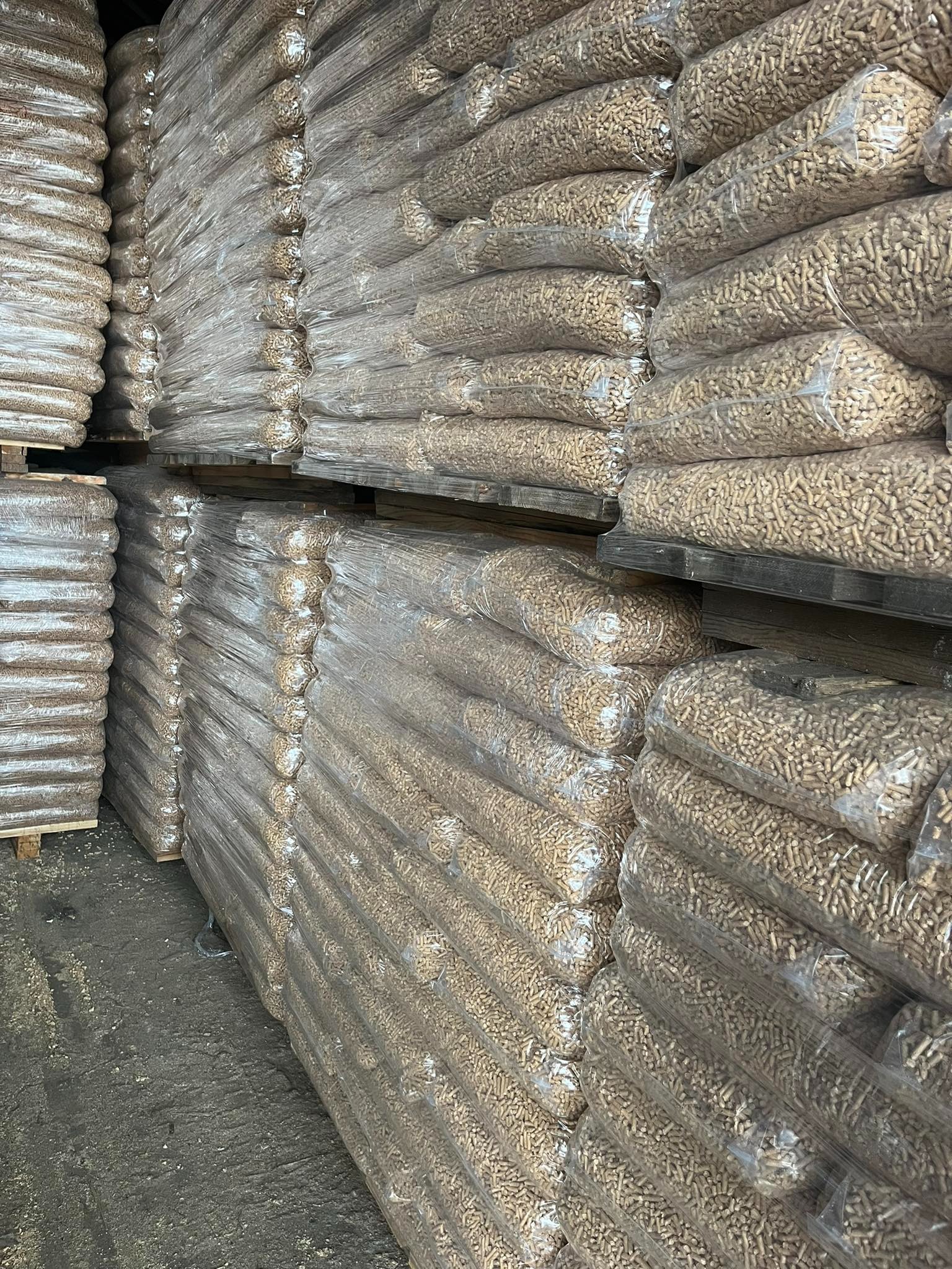 Magazynowane worki z pelletem drzewnym ułożone na drewnianych paletach, owinięte folią, widoczne nierówności betonu na podłodze.