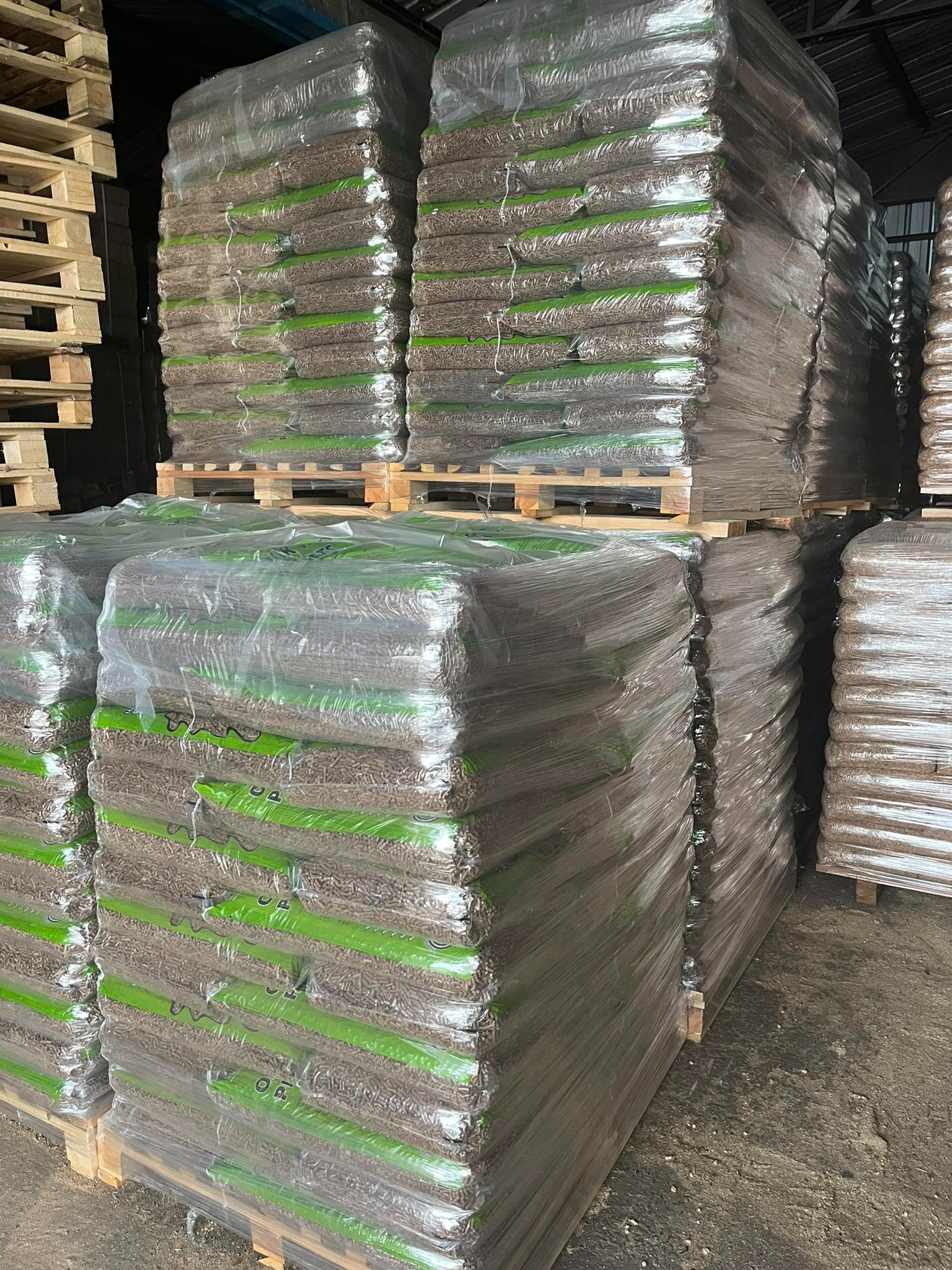 Ułożone w stosy palety z workami z pelletem, owinięte folią stretch, widoczne w magazynie.