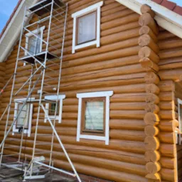 Drewniany dom z bali w trakcie malowania elewacji, widoczne rusztowanie i pomalowane fragmenty ściany na jasny odcień brązu.