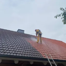 Pracownik w kombinezonie i kasku, zabezpieczony linami, czyści dach z czerwonej dachówki myjką ciśnieniową; widoczna różnica między umytą a brudną częścią dachu, dom z bali i drabina oparta o dach.