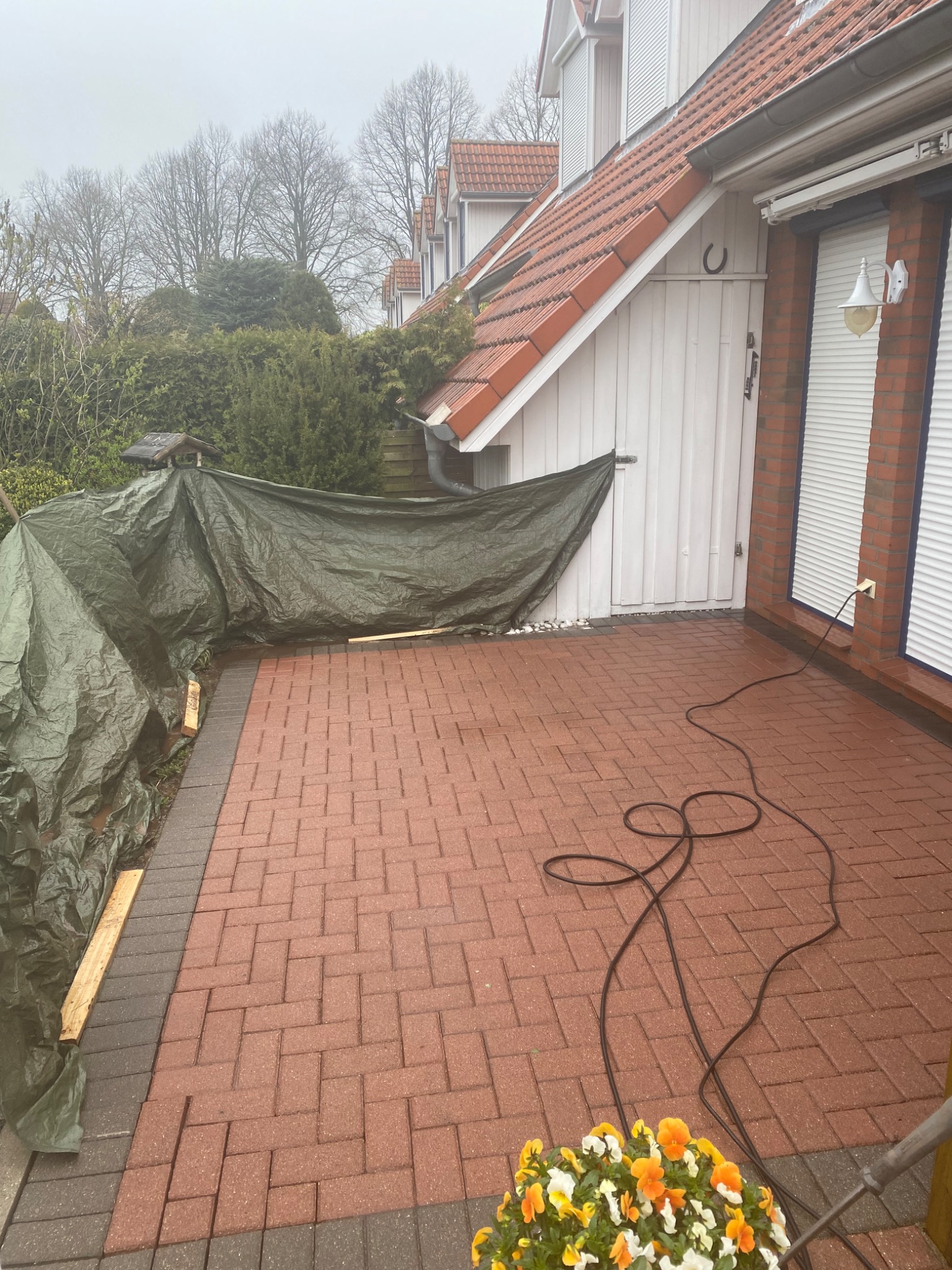 Częściowo ułożona czerwona kostka brukowa przed domem, zabezpieczona plandeką, z widocznym przewodem i kwiatami w doniczce.