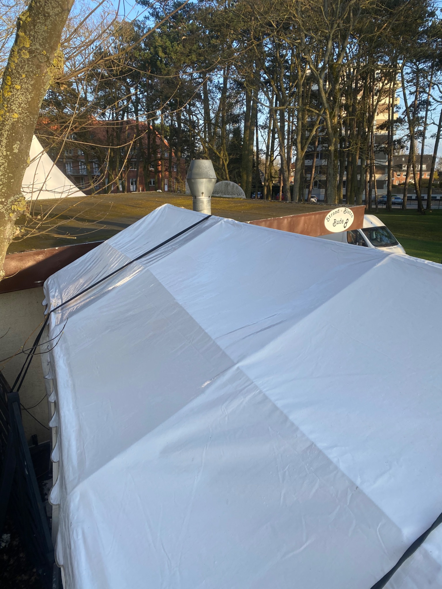 Biały namiot ochronny zakrywający dach budynku z kominem, widoczny napis 'Strand-Bistro Bude 8' oraz samochód dostawczy w tle, drzewa i budynki w oddali.