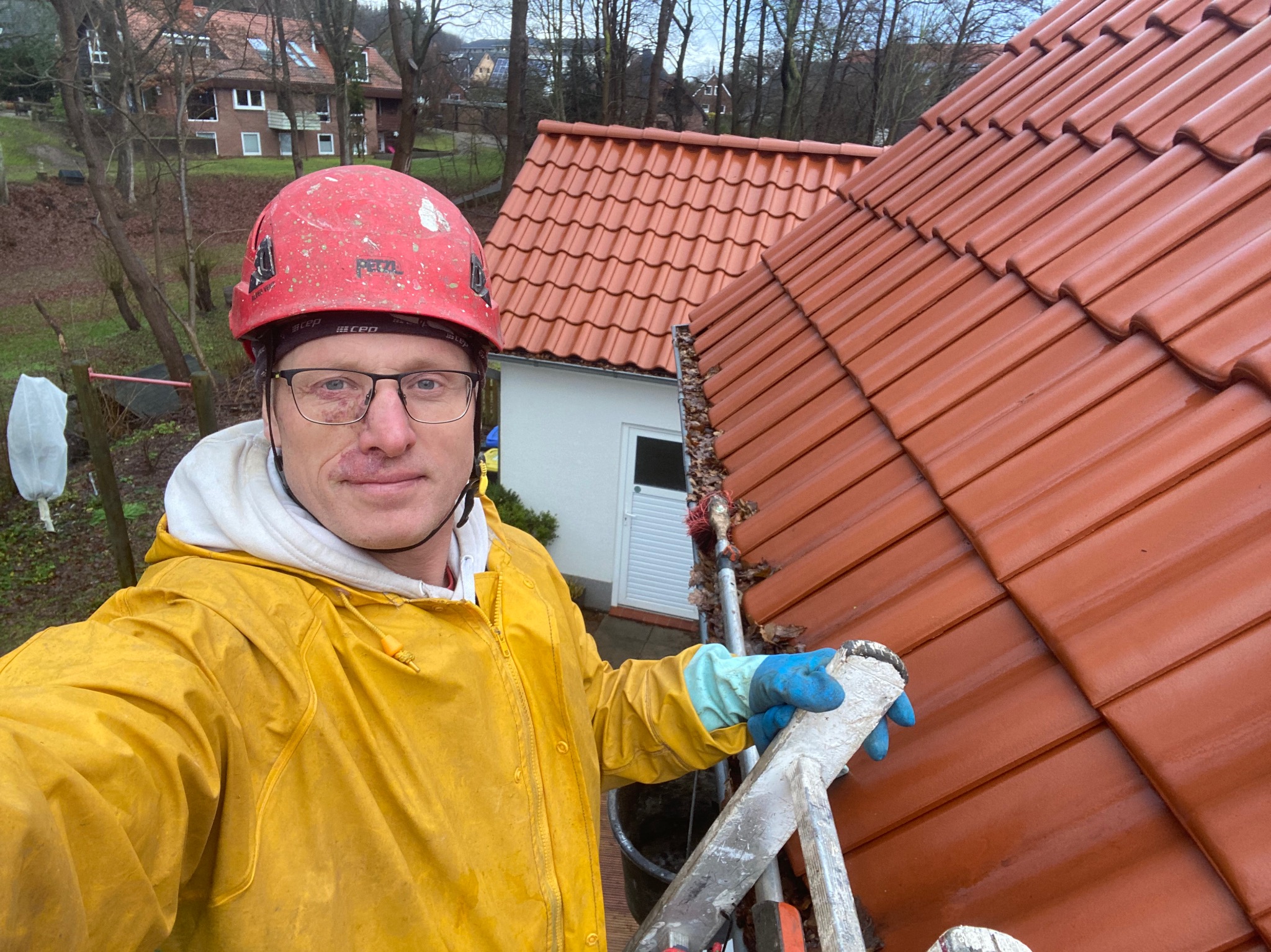 Pracownik w żółtej kurtce i czerwonym kasku, z lekkimi zabrudzeniami na twarzy, stoi na drabinie na tle dachu z czerwonej dachówki podczas pochmurnej pogody.