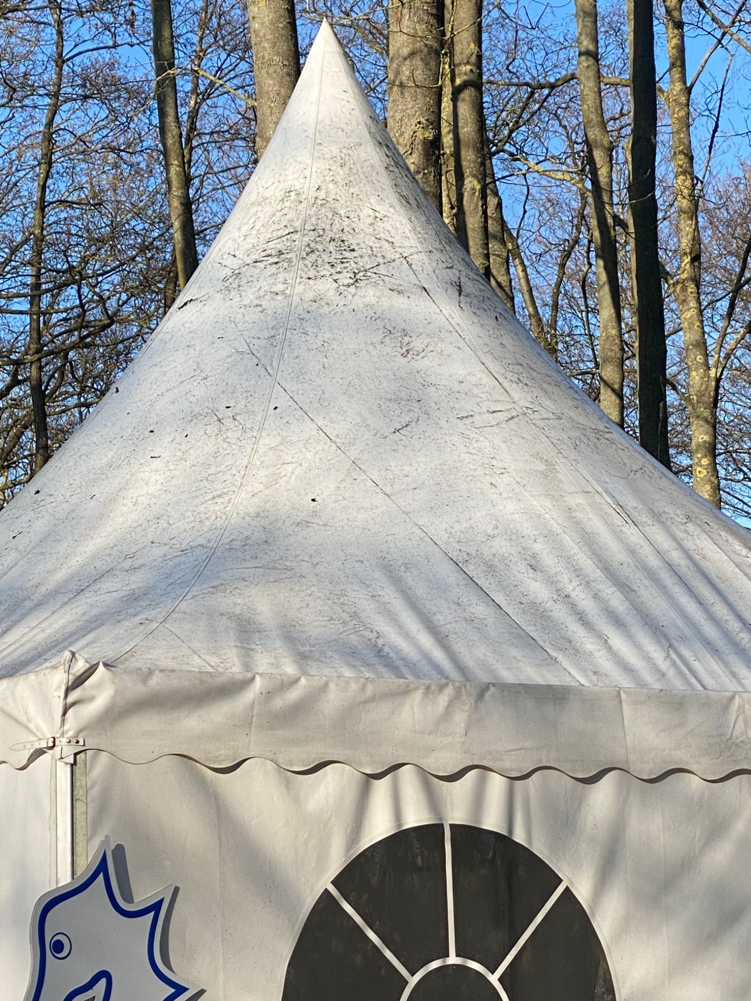 Biały, stożkowy namiot imprezowy z widocznymi zabrudzeniami na poszyciu, okrągłym oknem i niebieskim logo na tle nagich drzew.