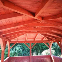 Wnętrze nowej, drewnianej altany w odcieniach czerwieni, z widokiem na zielony ogród przez balustradę.